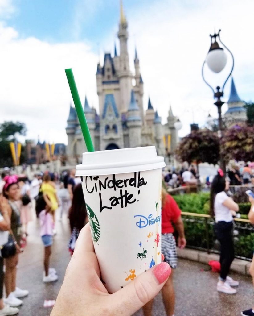 Image of Cinderella latte, a Starbucks drink listed on the Magic Kingdom Bucket list. Beverage is being held in front of Cinderella's castle at Magic Kingdom in Disney World.